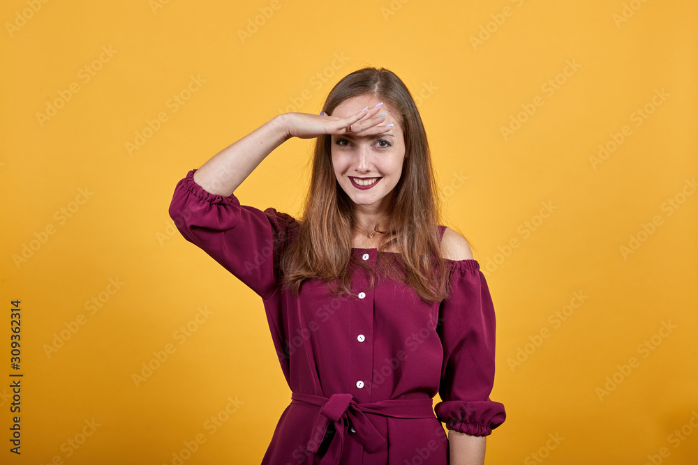 Young lady in burgundy bluse with bow over isolated orange background looking far away with hand to see something