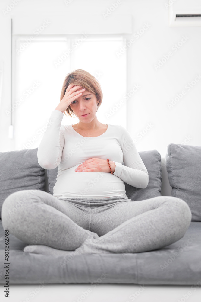 Fototapeta premium Pregnant tired exhausted woman with stomach and head issues at home on a couch, being sick.