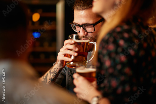 Handsome caucasian bearded tattooed man standing at pub with his friends and drinking beer.