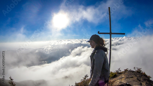A young woman standing on the top of volcano Inierie in Bajawa, Flores, Indonesia, next to a cross. She is surrounded by thick clouds. Sun tries to get through the thick layer of overcast. Freedom photo