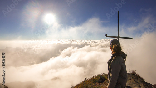A young woman standing on the top of volcano Inierie in Bajawa, Flores, Indonesia, next to a cross. She is surrounded by thick clouds. Sun tries to get through the thick layer of overcast. Freedom photo