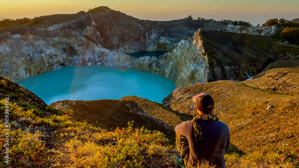 A man watching the sunrise over the Kelimutu volcanic crater lakes in Moni, Flores, Indonesia. Skyline is orange, sun slowly rising. Man is relaxed and calm, enjoying the view on turquoise lake