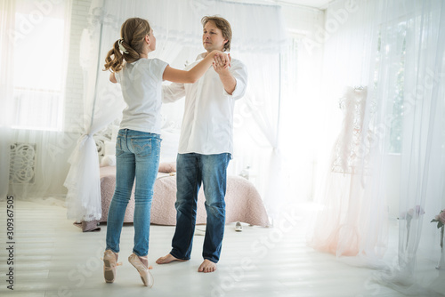 Young proud dad is dancing with his teenage daughter a ballerina in pointe shoes and jeans standing in a chic room with a beautiful interior. Concept of good relationship between father and daughter
