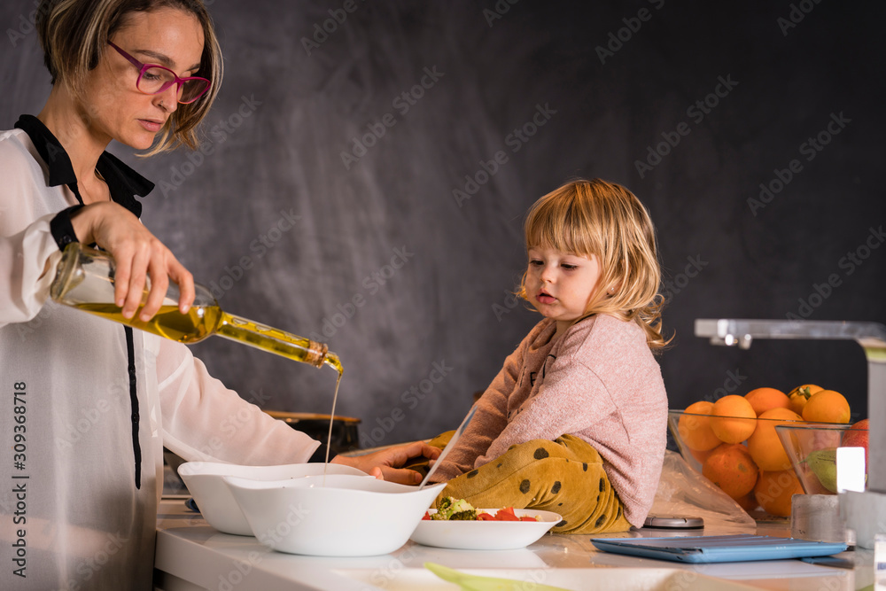 Woman dressing salad with a little girl sitting next to her in the kitchen