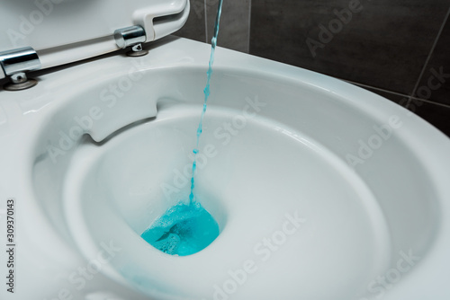 close up view of blue liquid detergent pouring into ceramic clean toilet bowl in modern restroom with grey tile