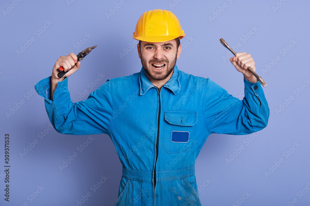 Close up portrait of angry construction worker engineer wearing yellow helmet and blue uniform, keeping hands up, shouting something with anger, bearded guy holding wrench tools with both hands. Close up portrait of angry construction worker engineer wearing yellow helmet and blue uniform, keeping hands up, shouting something with anger, bearded guy holding wrench tools with both hands.