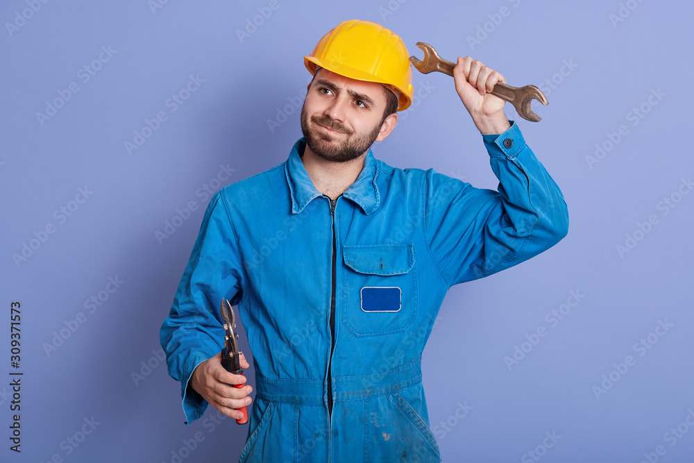 Young construction worker holding wrench tools in hands and looking aside with thoughtful facial expression, posing against blue studio background, bearded male thinks how to repair something. Young construction worker holding wrench tools in hands and looking aside with thoughtful facial expression, posing against blue studio background, bearded male thinks how to repair something.