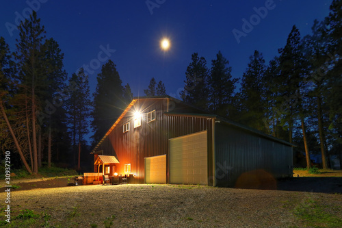 Obraz na plátně Large metal barn at night with fire at round table and outdoor furniture