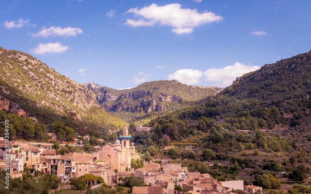 Valldemossa church and surrounding town with mountains, forests and blue sky with white clouds, Mallorca, Spain.