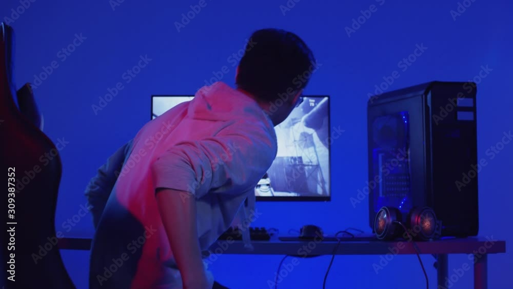 Rear view of young Asian man wearing grey hoody sitting down on chair at computer and starting playing online video game Rear view of young Asian man wearing grey hoody sitting down on chair at computer and starting playing online video game