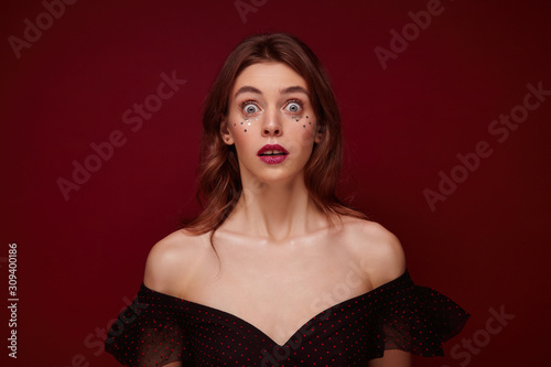 Open-eyed young pretty female with brown wavy hair wearing festive evening makeup while standing over claret background, rounding amazedly eyes and looking at camera with shocked face photo