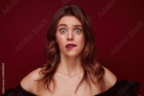 Indoor photo of confused young brunette woman with wavy hairstyle and silver little stars on her face looking suprisedly at camera and biting underlip while posing against claret background