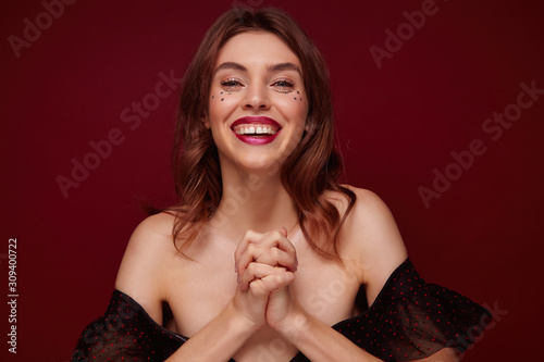 Portrait of positive young lovely brown haired female dressed in festive clothes looking happily at camera and smiling widely, crossing raised arms while standing against claret background photo