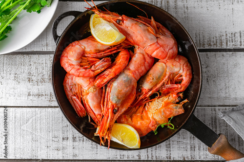 Jumbo prawns with lemon wedges served on cast iron pan on white wooden background, closeup view