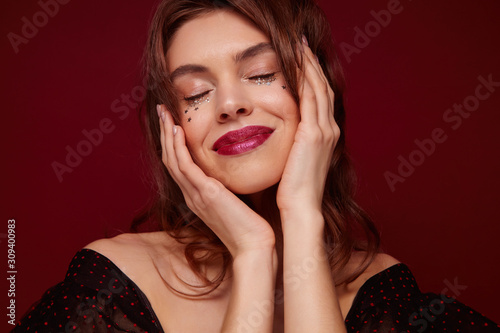 Enjoyable young brown haired pretty woman with red lips and little silver stars on her face smiling pleasantly with closed eyes, holding face with raised hands while posing against claret background