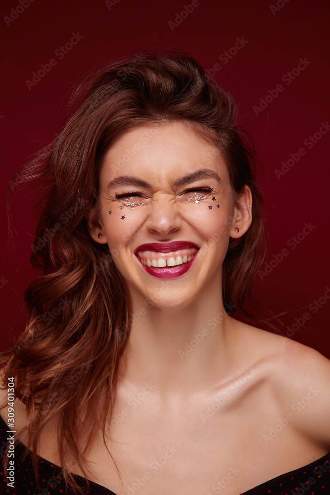 Beautiful cheerful young lady with brown wavy hair laughing happily and showing her white perfect teeth, wearing evening makeup and small silver stars on her face, isolated over claret background Beautiful cheerful young lady with brown wavy hair laughing happily and showing her white perfect teeth, wearing evening makeup and small silver stars on her face, isolated over claret background
