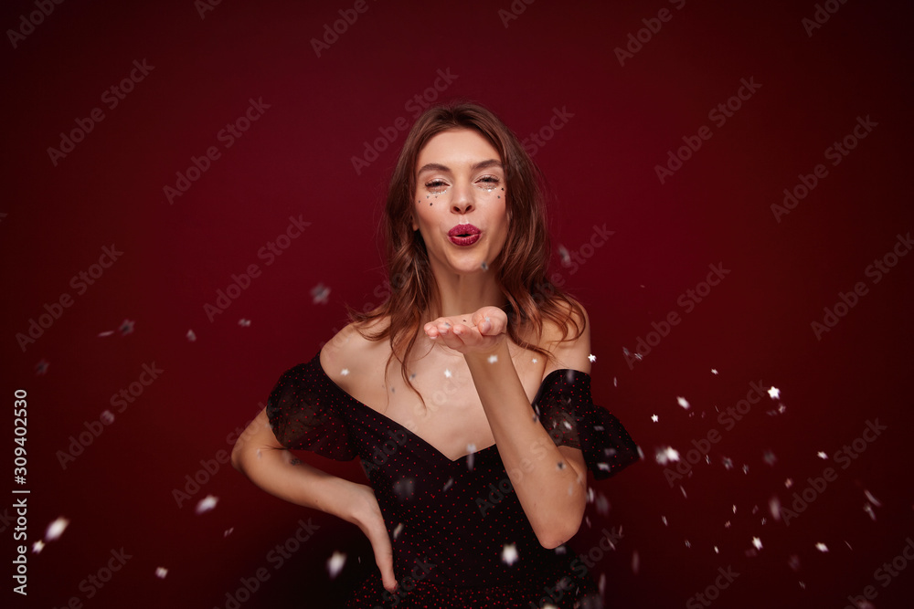 Attractive young brunette lady with wavy hairstyle wearing festive clothes while standing over claret background, having cheerful moments in her life during new year party