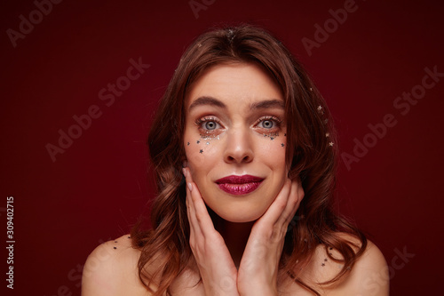 Close-up of beautiful young female with brown wavy hair wearing evening makeup while posing over claret background, looking calmly at camera and touching gently her face