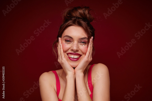 Indoor photo of attractive cheerful young female with bun hairstyle and festive makeup holding her face with palms and smiling widely at camera, standing over burgundy background in top with straps