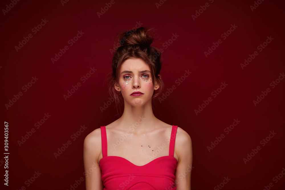 Portrait of unhappy attractive young female with festive makeup wearing her brown hair in bun, looking at camera with sad face and keeping lips folded, posing over burgundy background with hands down