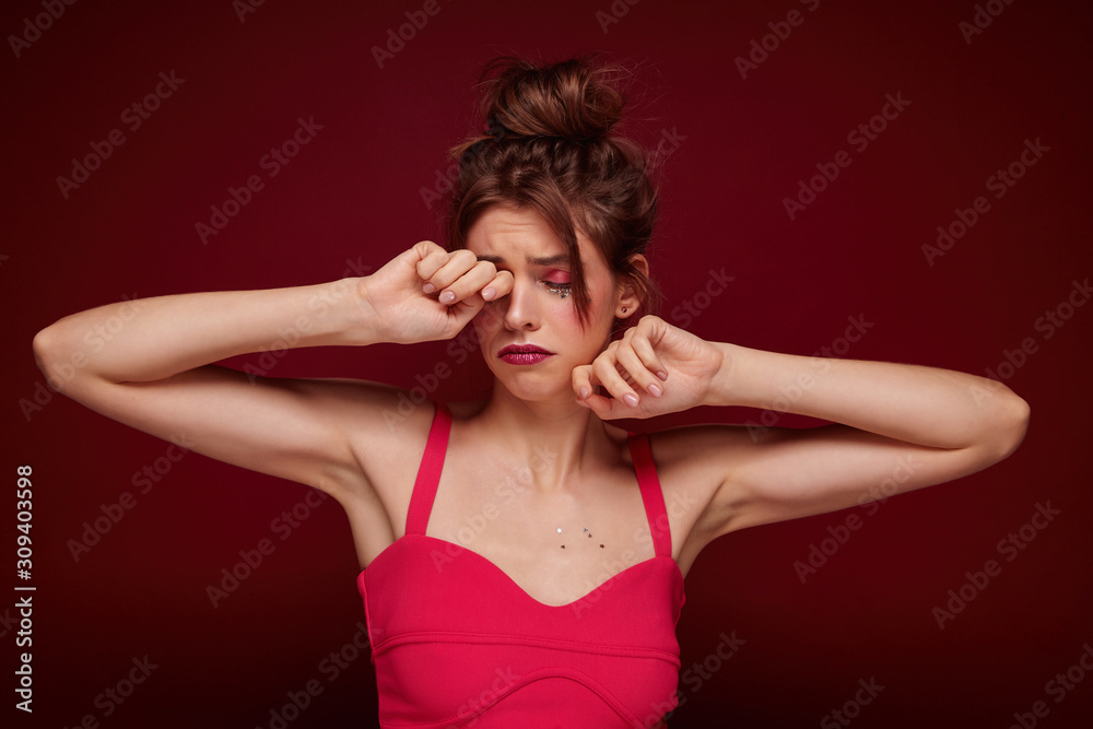 Tired young pretty brunette woman with bun hairstyle raising hands to her face and rubbing her eye wearily, dressed in pink top with straps while standing over burgundy background