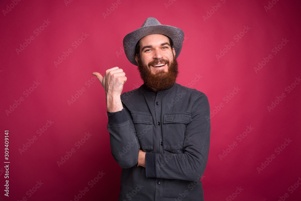 Cheerful young bearded man wearing grey hat and pointing away at copyspace Cheerful young bearded man wearing grey hat and pointing away at copyspace