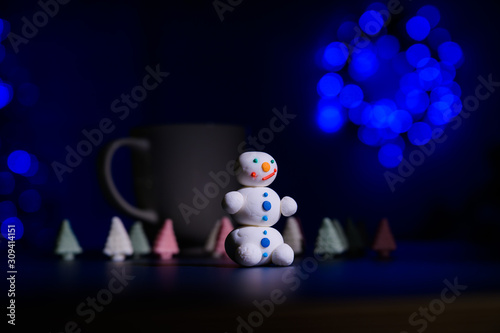 Cute snowman and little Christmas trees made of marshmallows on the background of Christmas lights. White mug of hot chocolate and sweets. photo