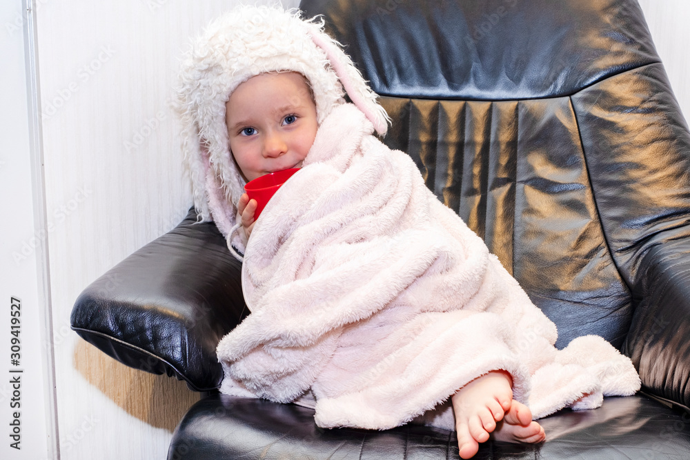 Girl in a winter hat with ears on a black armchair. A child in a warm bathrobe drinks from a red mug Girl in a winter hat with ears on a black armchair. A child in a warm bathrobe drinks from a red mug