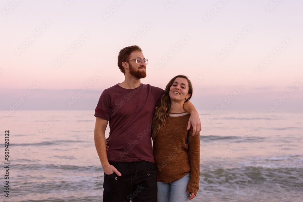 Young caucasian couple hugging and laughing together at a sunrise on the beach, redhead boy and blonde girl