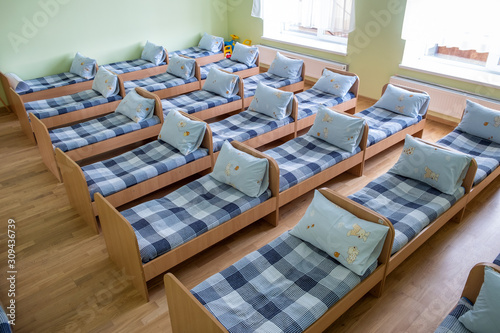 Many small beds with fresh linen in daycare preschool empty bedroom interior for comfortable afternoon nap of the kids. © bilanol