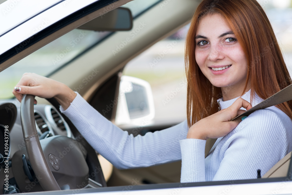 Wide angle view of young redhead woman driver fastened by seatbelt driving a car smiling happily.