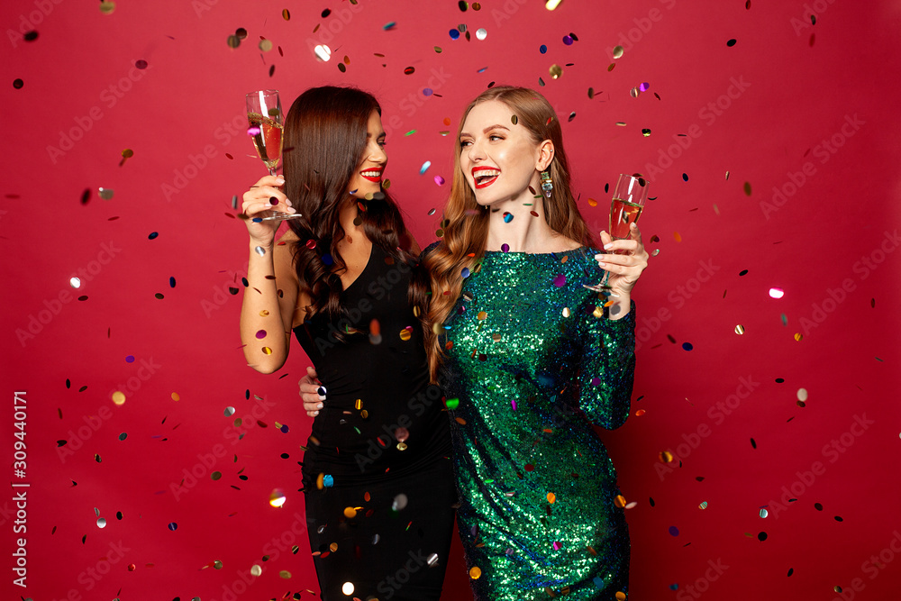 Plakat two beautiful models, a redhead and a brunette in New Year's dresses, having fun and smiling with glasses of champagne, confetti flying around on a red background. New Year or Christmas photo