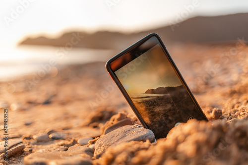 The concept of the photos on the phone. The smartphone lies buried in the sand on the beach, and takes a photo of the beach in the background. Copy space photo