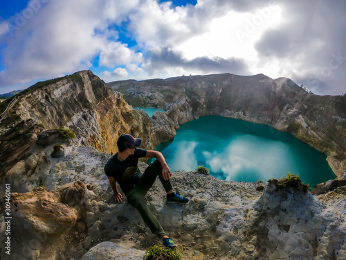 Man sitting at the volcano rim and watching the Kelimutu volcanic crater lakes in Moni  Flores  Indonesia. Man is relaxed and calm  enjoying the view on lake shining with many shades of turquoise