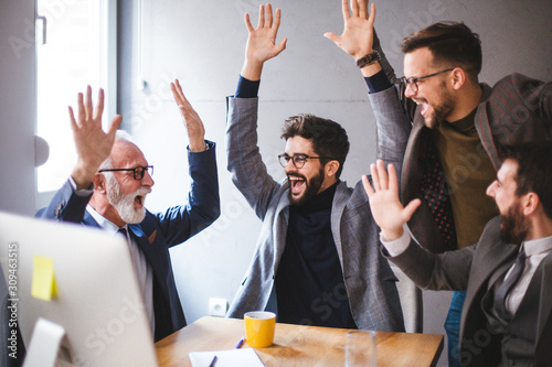 Four Caucasian bearded managers happy and pleasantly surprised with their new business opportunities, laughing and raising their hands in the air.