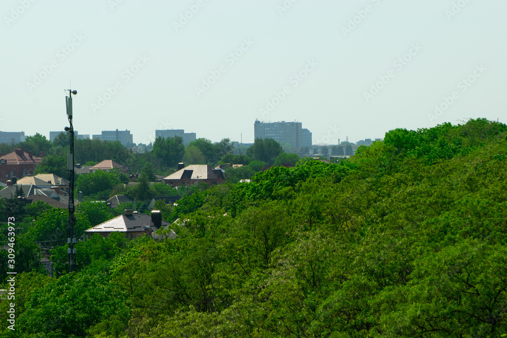 The repeater tower rises above the green crowns of trees against the background of houses in the distance