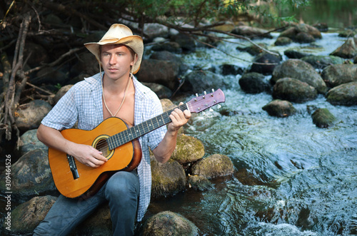 Handsome guy in cowboy hat playing the guitar on rocky river shore photo