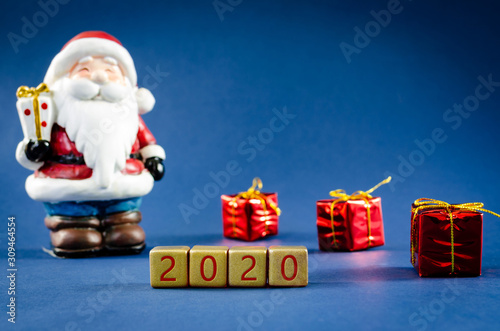 Blue christmas background with Santa Clause figure and gifts in a conceptual image with 2020 spelled on gold blocks.