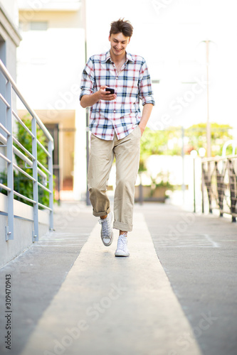 Full length happy young man walking with mobile phone outside