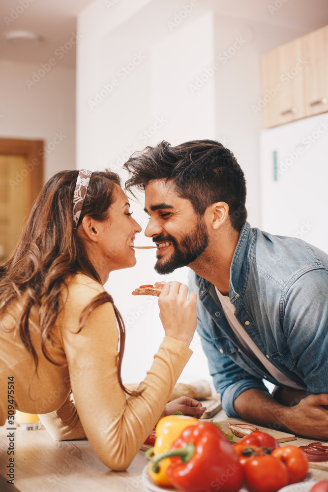 A man and a woman moments before kissing over the kitchen countertop. The woman is holding a carrot. Vegetables are layed on the counter as well.