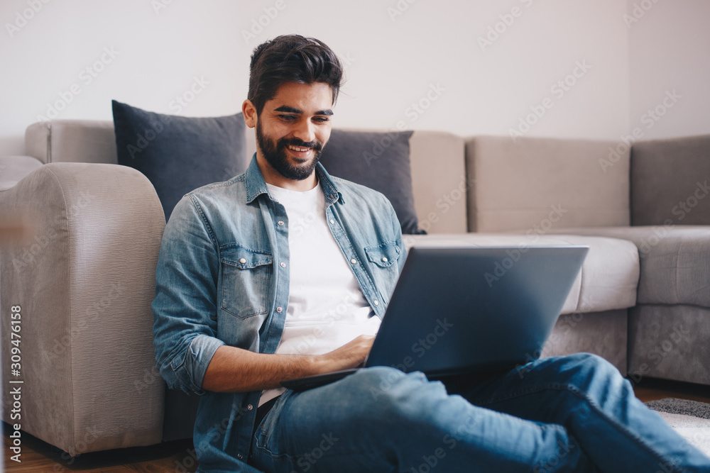 A young man chilling in his livingroom with a laptop on his lap. He is sitting on the floor, by a stylish couch. A young man chilling in his livingroom with a laptop on his lap. He is sitting on the floor, by a stylish couch.
