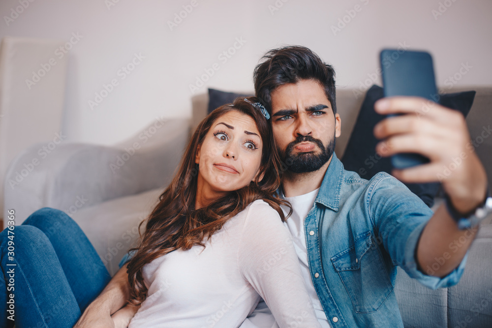 Fototapeta premium Cute Caucasian couple making funny faces while they are posing for a selfie with smart-phone and sitting by the couch in apartment.