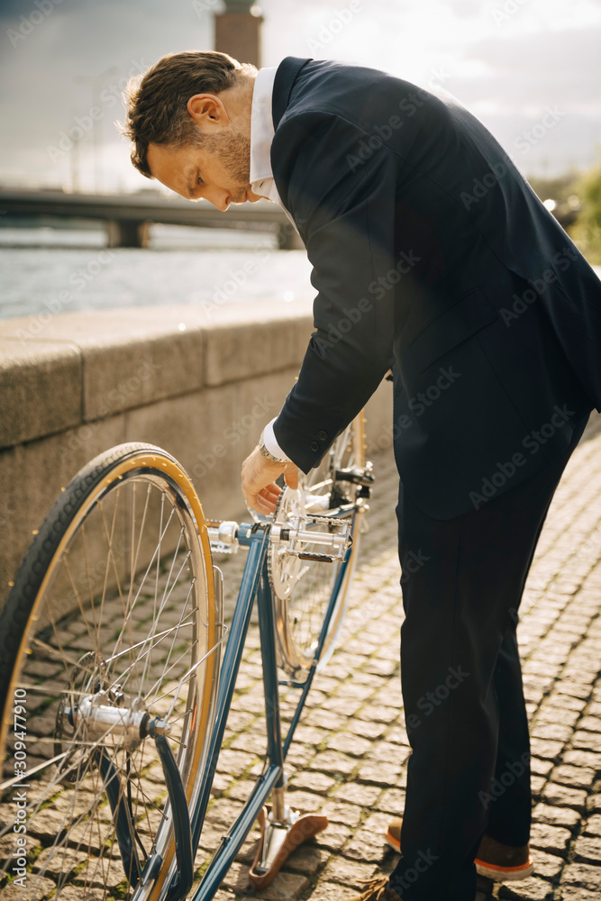 Side view of businessman repairing bicycle on footpath in city during sunny day