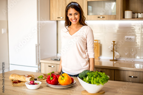 Young beautiful Caucasian brunette looking at camera  smiling while she s standing by the kitchen counter with vegetables and salads on it.