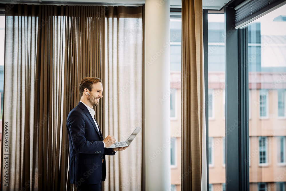 Smiling male legal professional talking through blue tooth while using laptop at office Smiling male legal professional talking through blue tooth while using laptop at office