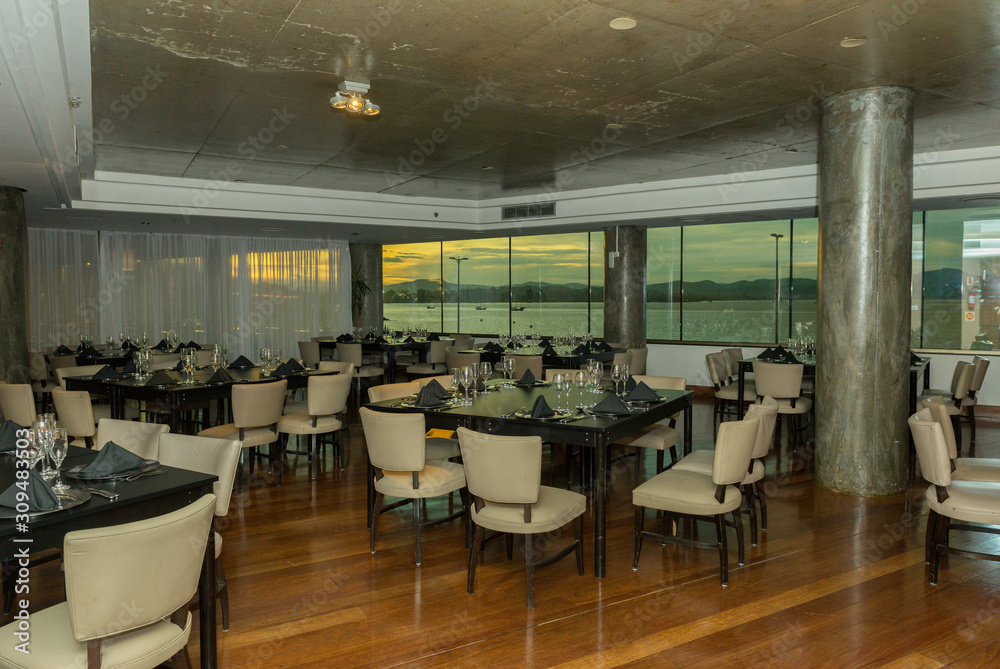 Luxurious party room with various tables, plates, cutlery and wine glasses. Windows overlooking the sea and sunset. Hall for events, parties, celebrations with beautiful view.