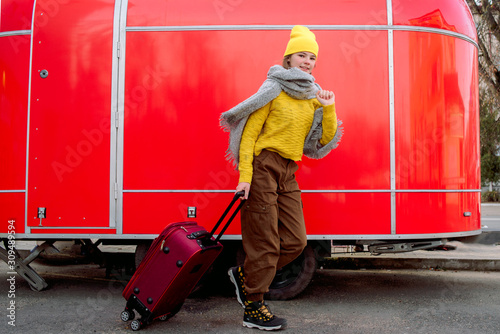 girl runs with a red suitcase on a red background. Large retro vintage van. Old car. Traveling in the winter. Girl in a yellow bright hat and knitted sweater. Travel concept.