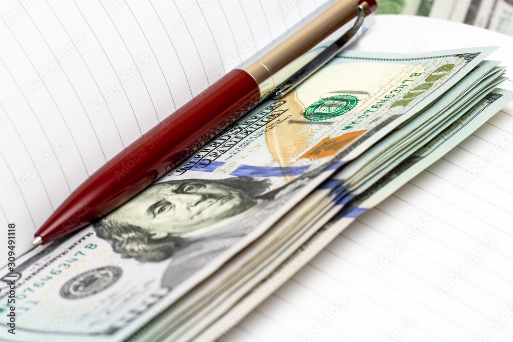 Close up of an empty business notepad and a pen with a pile of US dollars Close up of an empty business notepad and a pen with a pile of US dollars
