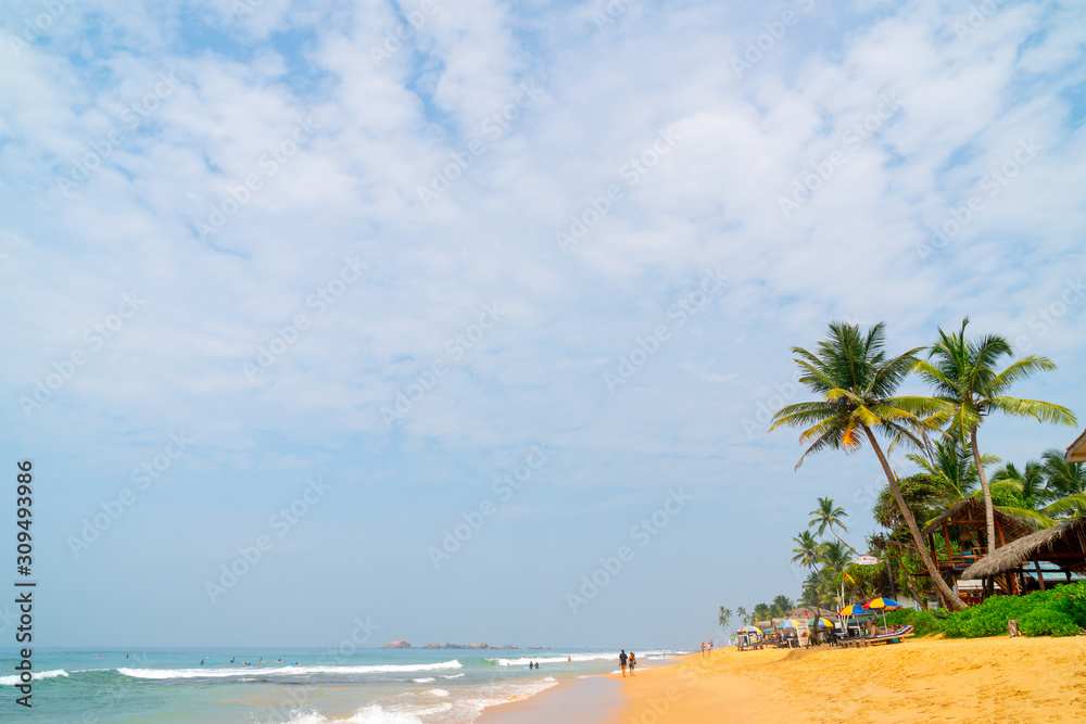 Hikkaduwa, Sri Lanka. March 8, 2018. Beach on the Indian Ocean. Sunny day, yellow sand, palm trees and foam waves.