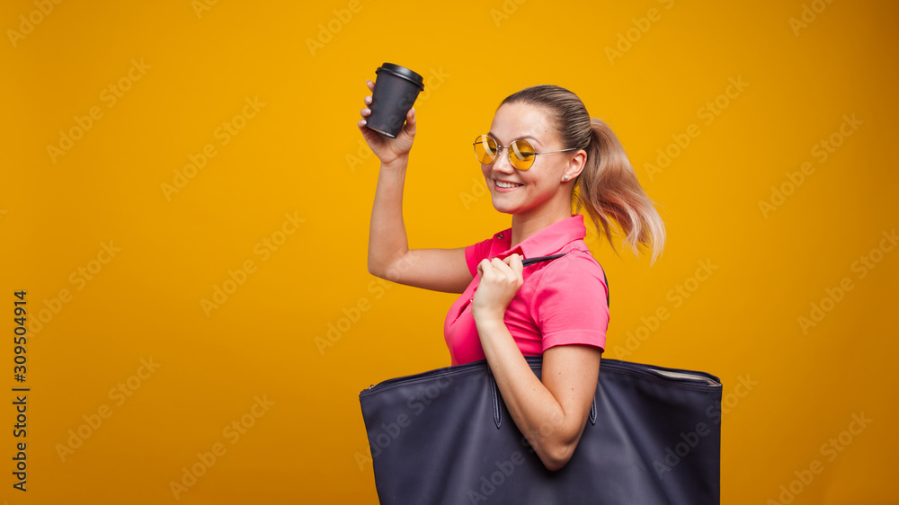 Cute blonde in sunglasses and t-shirt with bag on shoulder, drinking her hot drink, portrait on yellow background Cute blonde in sunglasses and t-shirt with bag on shoulder, drinking her hot drink, portrait on yellow background